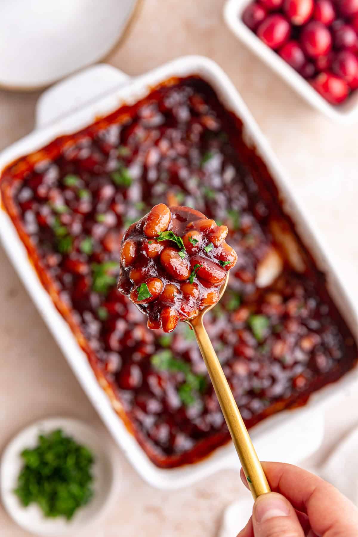 A dish of freshly baked cranberry bbq baked beans, garnished with parsley, with a spoon scooping some out.