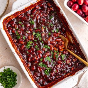 A dish of freshly baked cranberry bbq baked beans, garnished with parsley, with a spoon scooping some out.