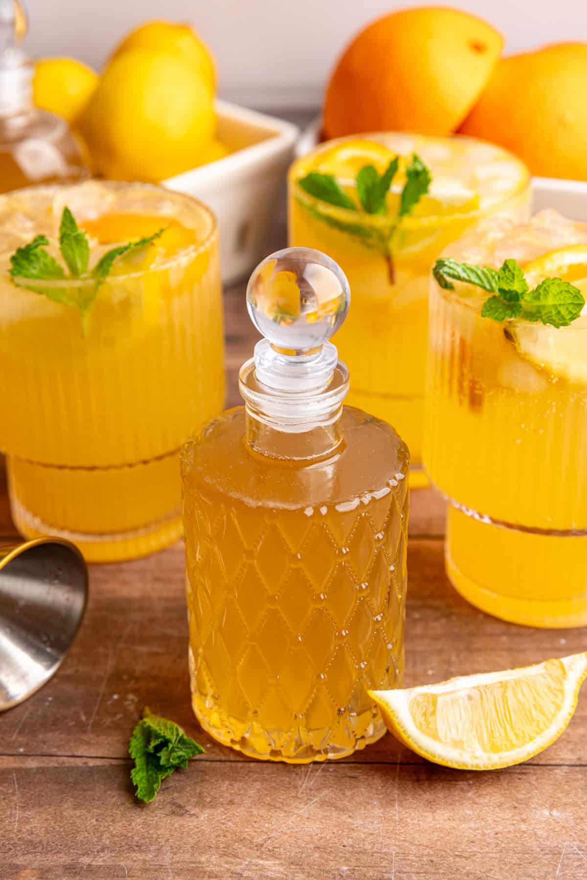 A bottle of lemon ginger syrup on a tabletop with glasses of citrus mocktail.