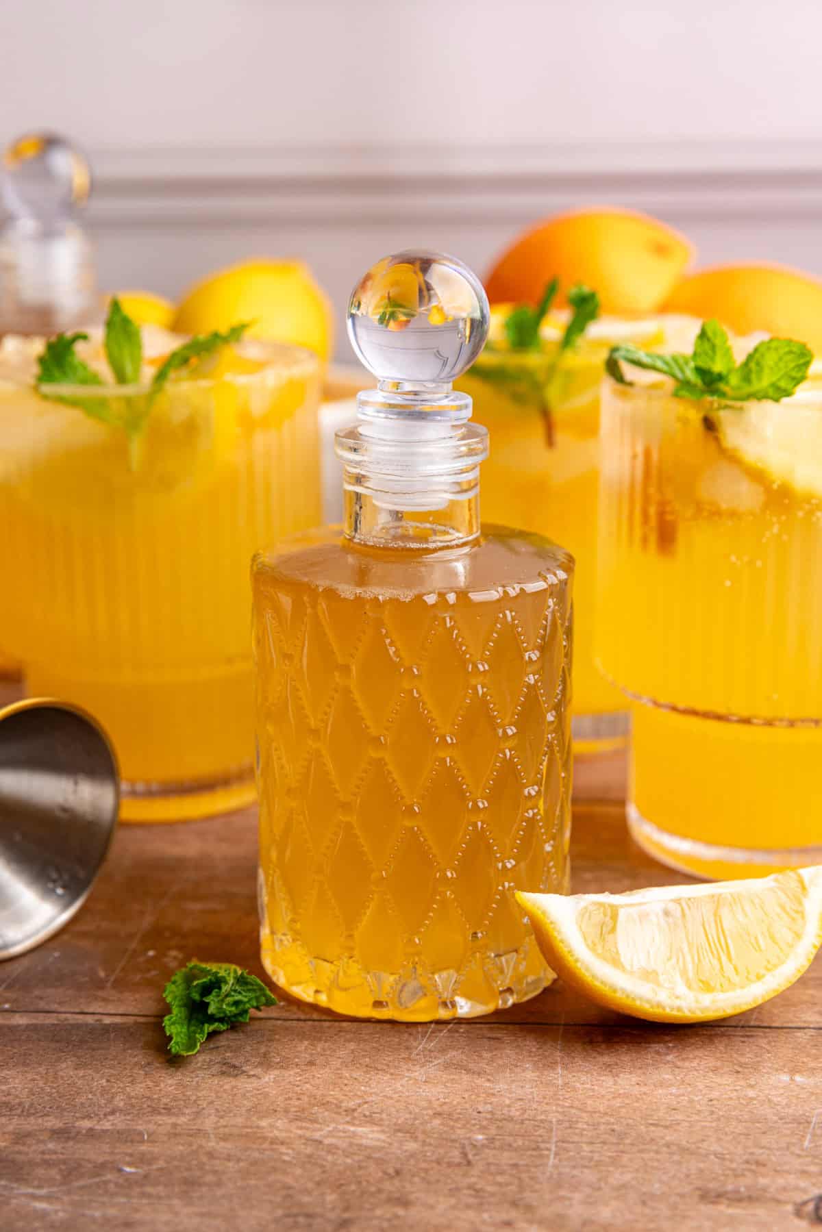 A bottle of lemon ginger syrup on a tabletop with glasses of citrus mocktail.
