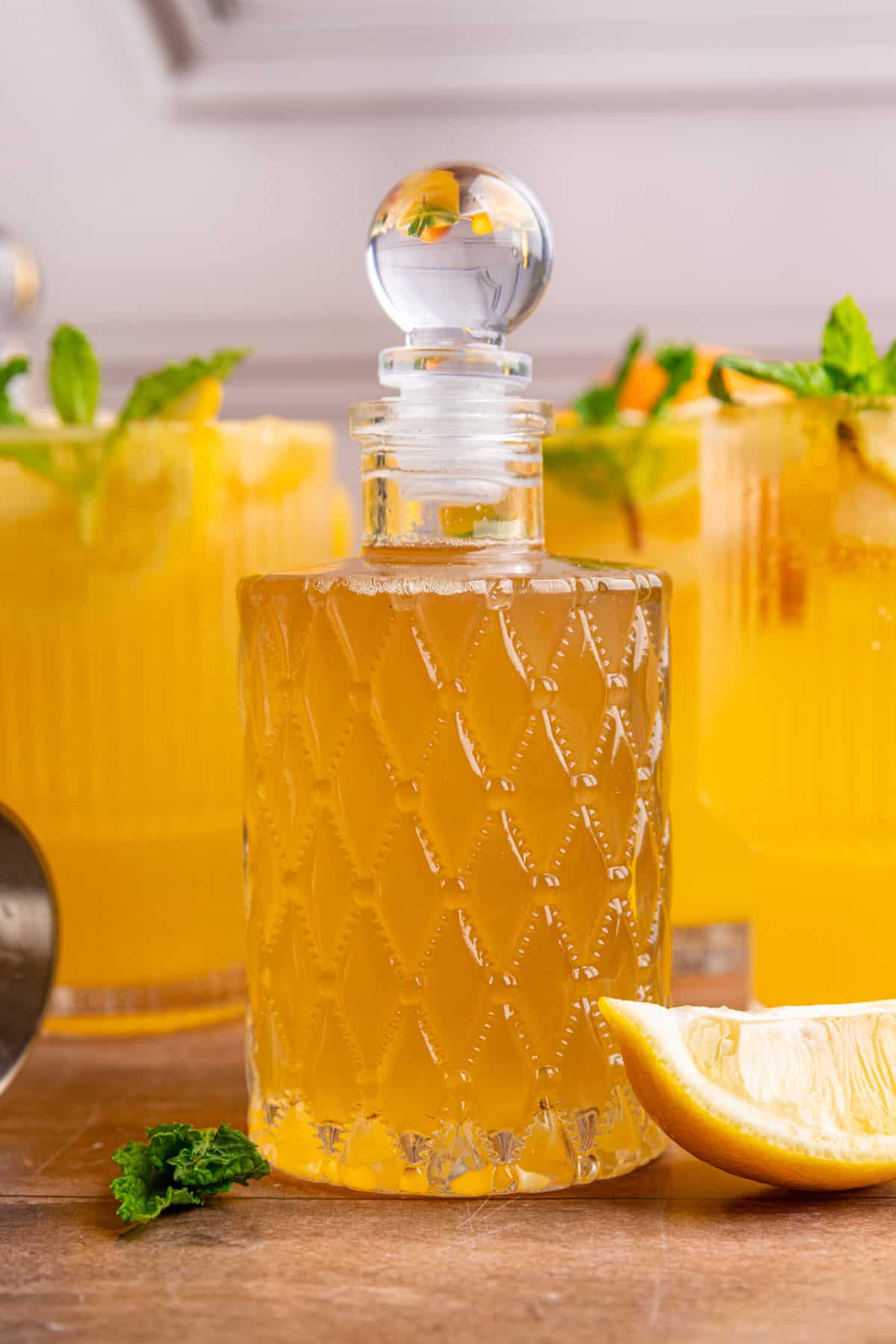 A bottle of lemon ginger syrup on a tabletop with glasses of citrus mocktail.