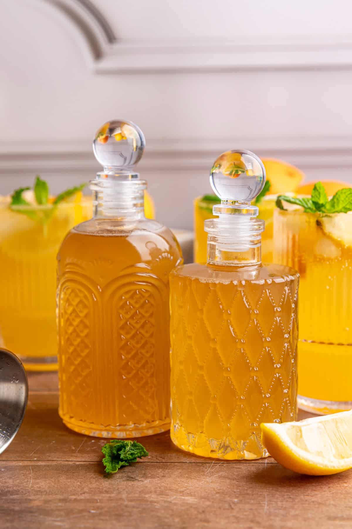 Two bottles of lemon ginger syrup on a tabletop with glasses of citrus mocktail.