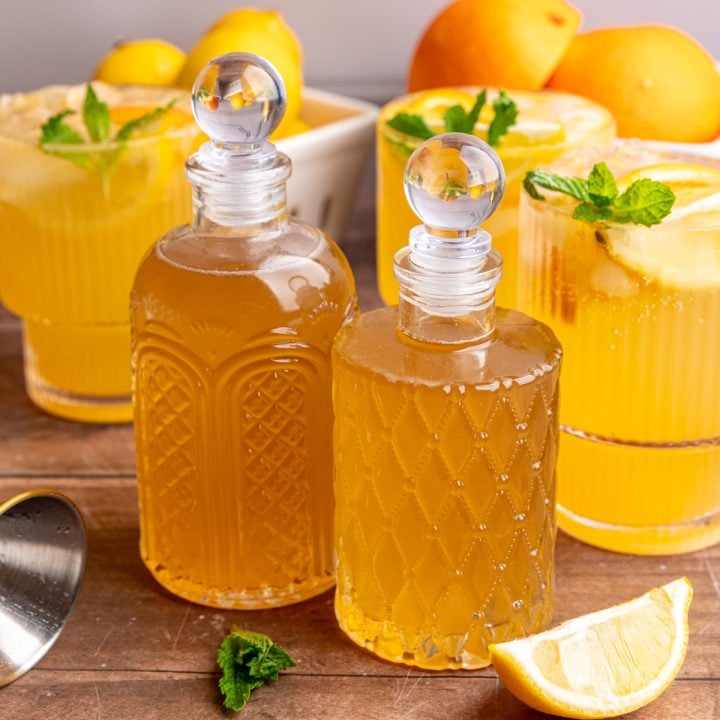 Two bottles of lemon ginger syrup on a tabletop with glasses of citrus mocktail.