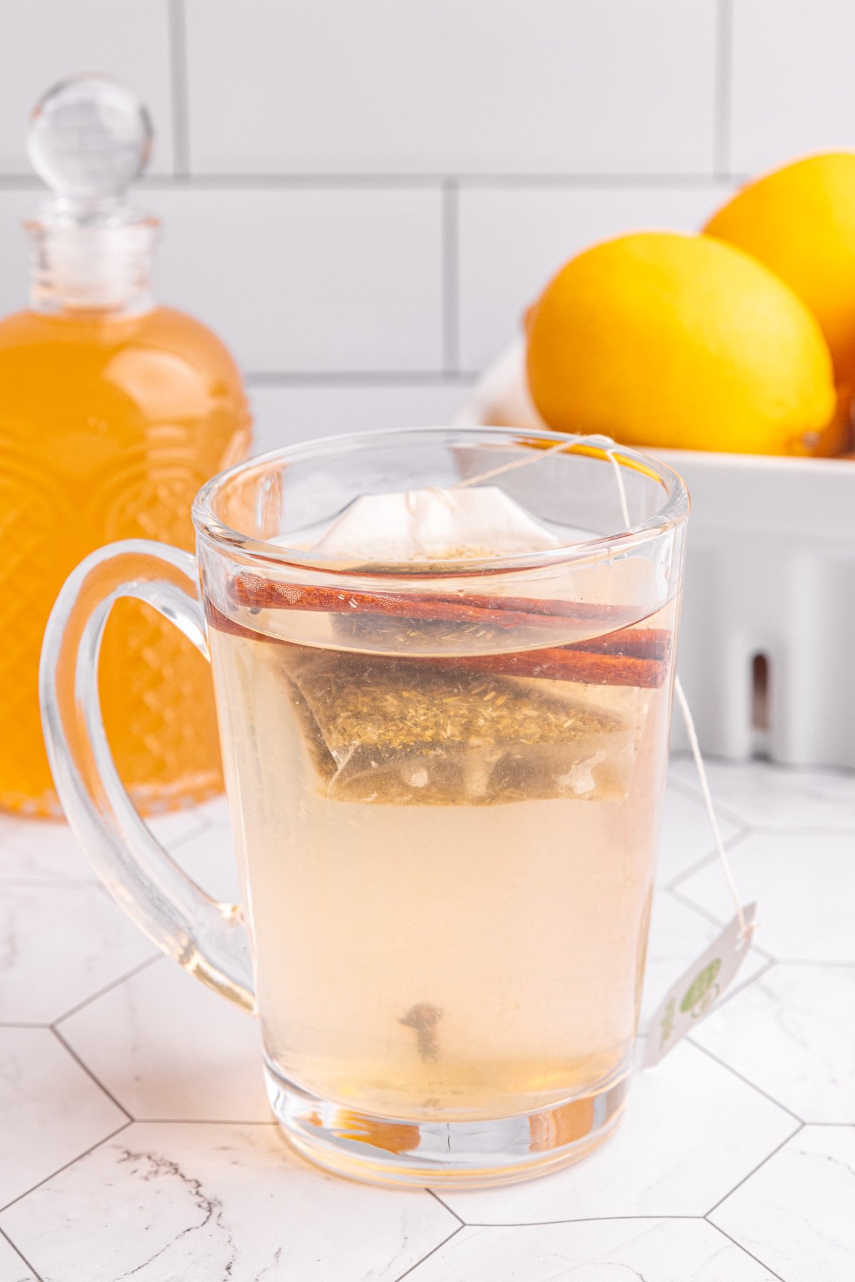 A glass of hot lemon ginger tea on a countertop, with a cinnamon stick and tea bag.