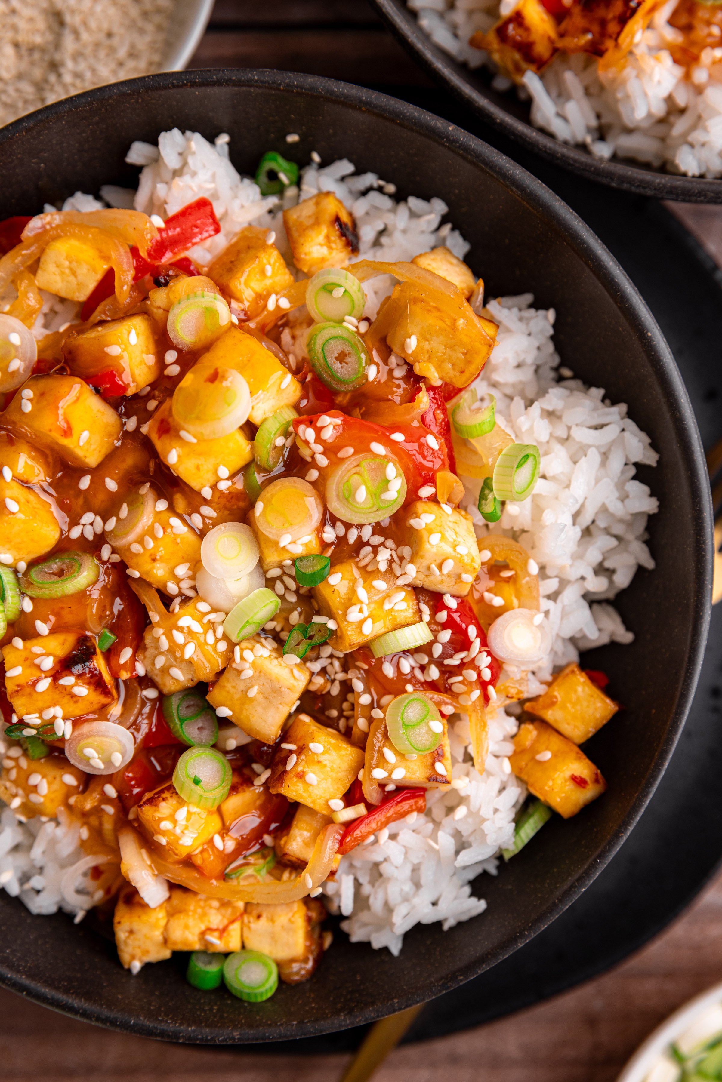 A bowl of sweet chili tofu, over rice, topped with scallions and sesame seeds.