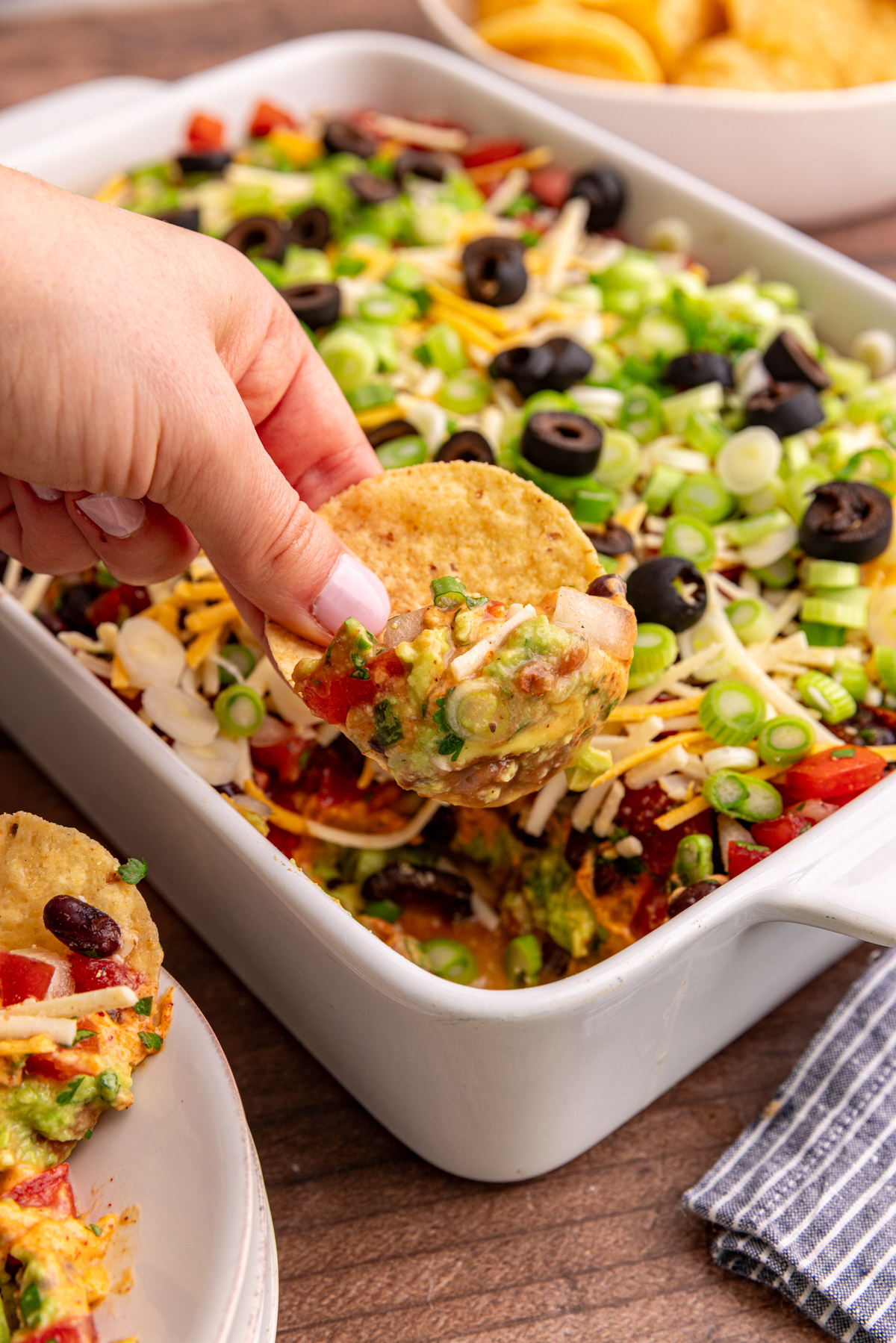 A hand scooping out a bite of vegan 7 layer dip with a tortilla chip.