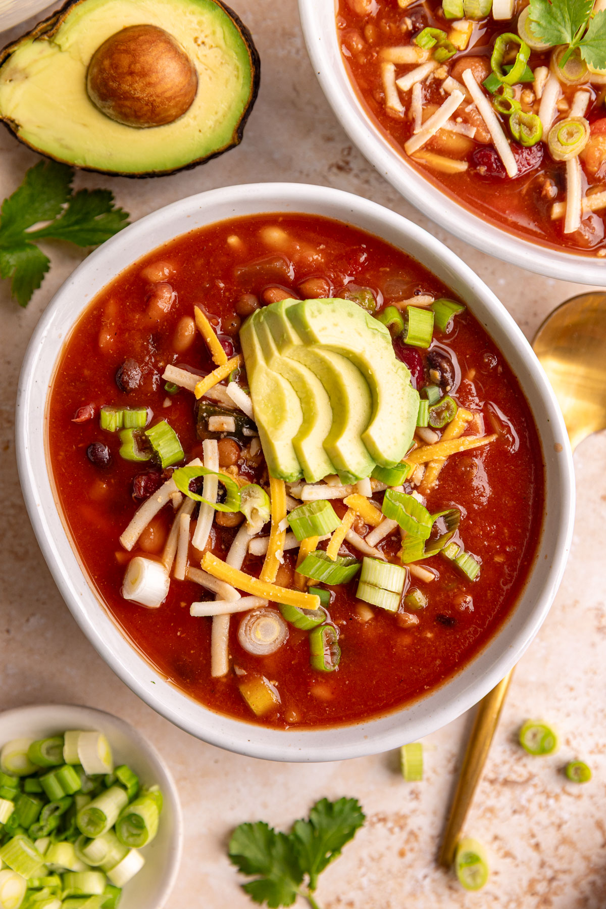 A bowl of vegan 5 bean chili garnished with avocado, vegan cheese, and scallions.