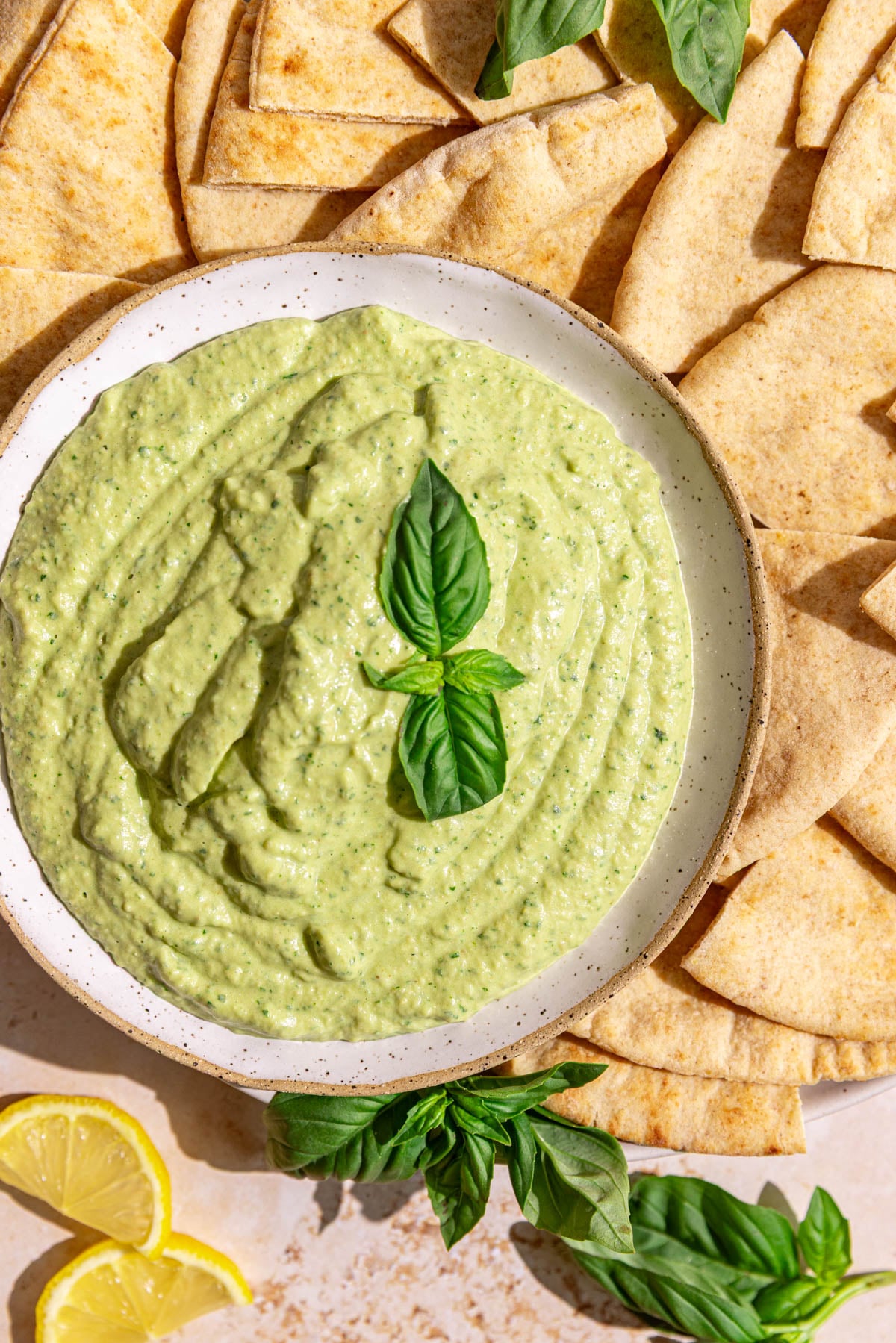 A bowl of basil hummus garnished with a fresh basil leaf, with a plate of pitas.