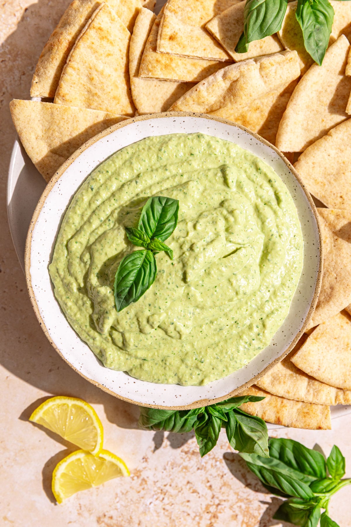 A bowl of basil hummus garnished with a fresh basil leaf, with a plate of pitas.