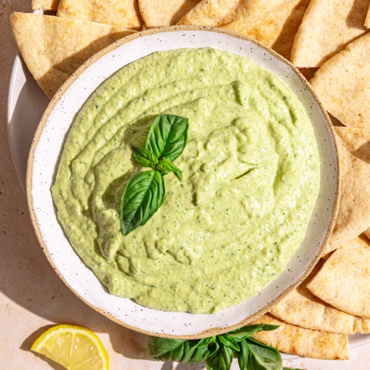 A bowl of basil hummus garnished with a fresh basil leaf, with a plate of pitas.