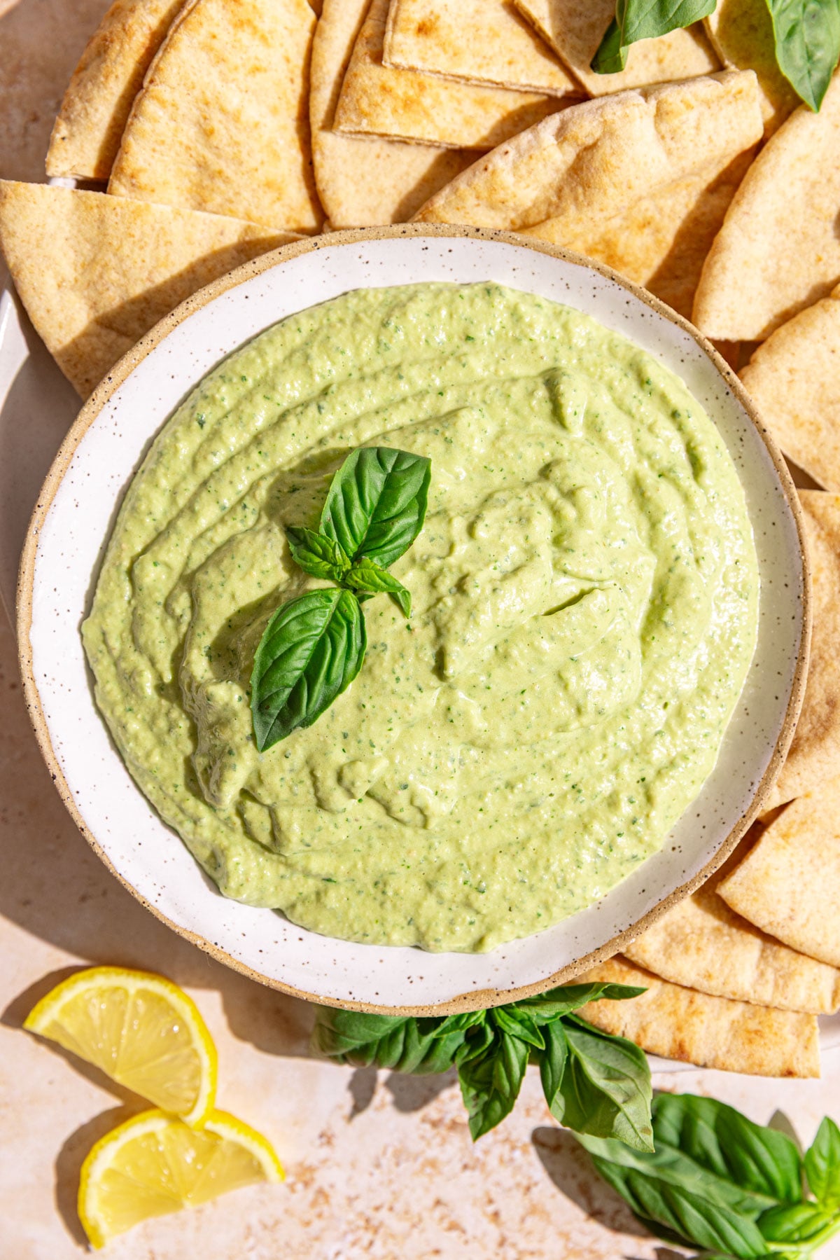 A bowl of basil hummus garnished with a fresh basil leaf, with a plate of pitas.