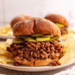 A lentil sloppy joe, with a layer of pickles, on a plate with potato chips.