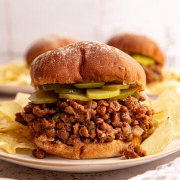 A lentil sloppy joe, with a layer of pickles, on a plate with potato chips.