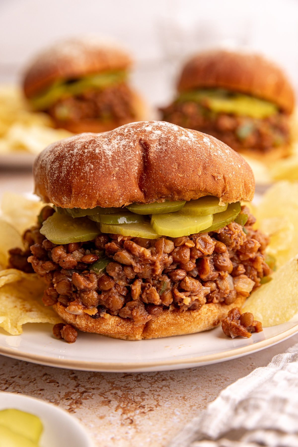 A lentil sloppy joe, with a layer of pickles, on a plate with potato chips.