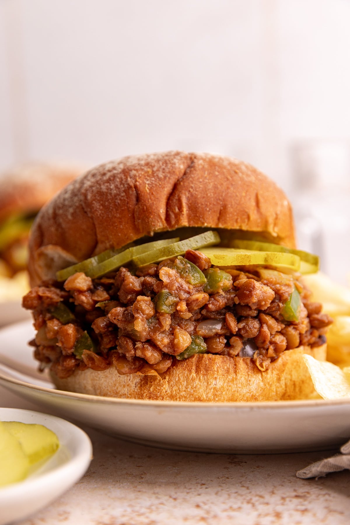 A lentil sloppy joe, with a layer of pickles, on a plate with potato chips.