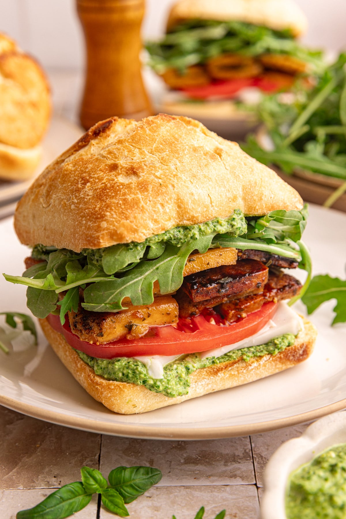 A plate with a vegan pesto sandwich, on a ciabatta roll, with pesto, tofu, and fresh veggies.