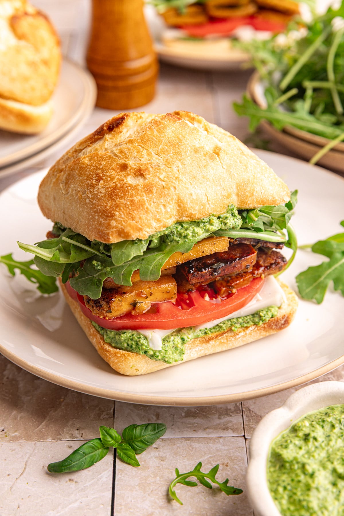 A plate with a vegan pesto sandwich, on a ciabatta roll, with pesto, tofu, and fresh veggies.