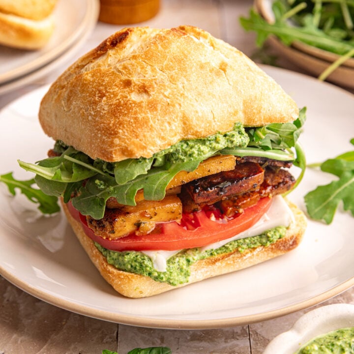 A plate with a vegan pesto sandwich, on a ciabatta roll, with pesto, tofu, and fresh veggies.
