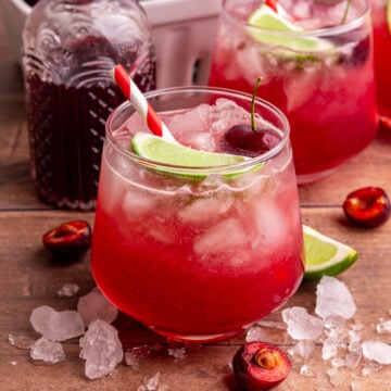 A cherry mocktail, on a table with ice, garnished with a lime wedge and a cherry.