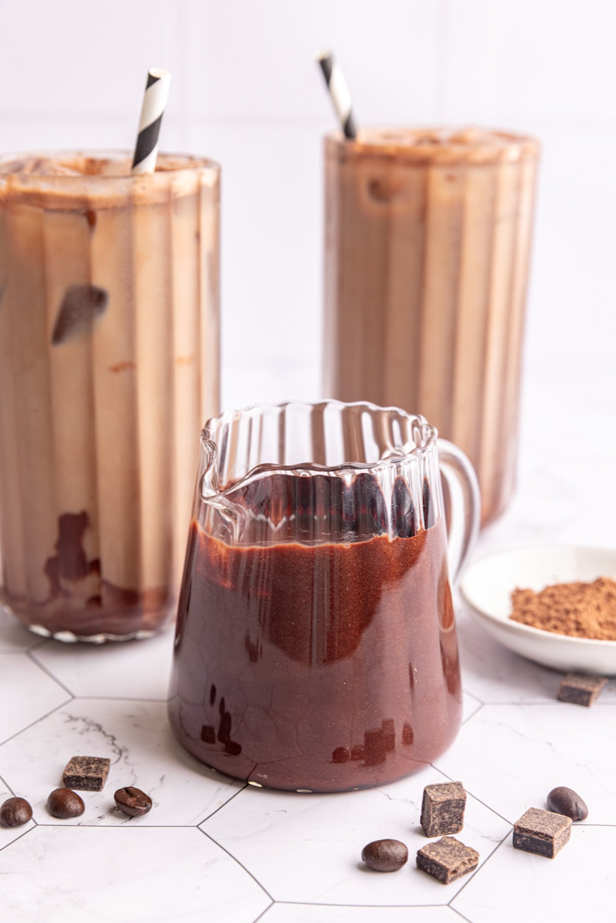 A small pitcher of vegan chocolate syrup next to two iced mochas.