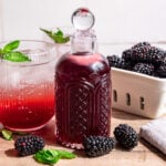 A bottle of blackberry shrub, next to a shrub mocktail and a berry basket of blackberries.