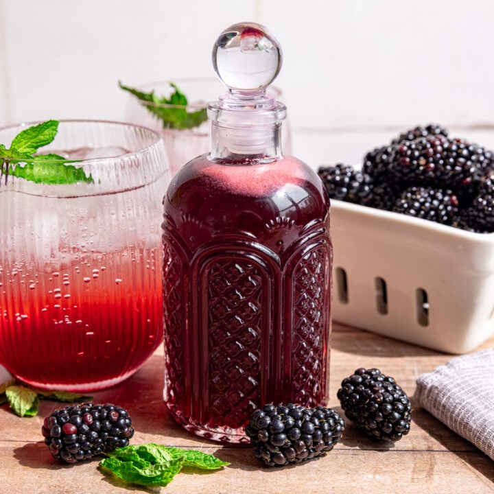 A bottle of blackberry shrub, next to a shrub mocktail and a berry basket of blackberries.