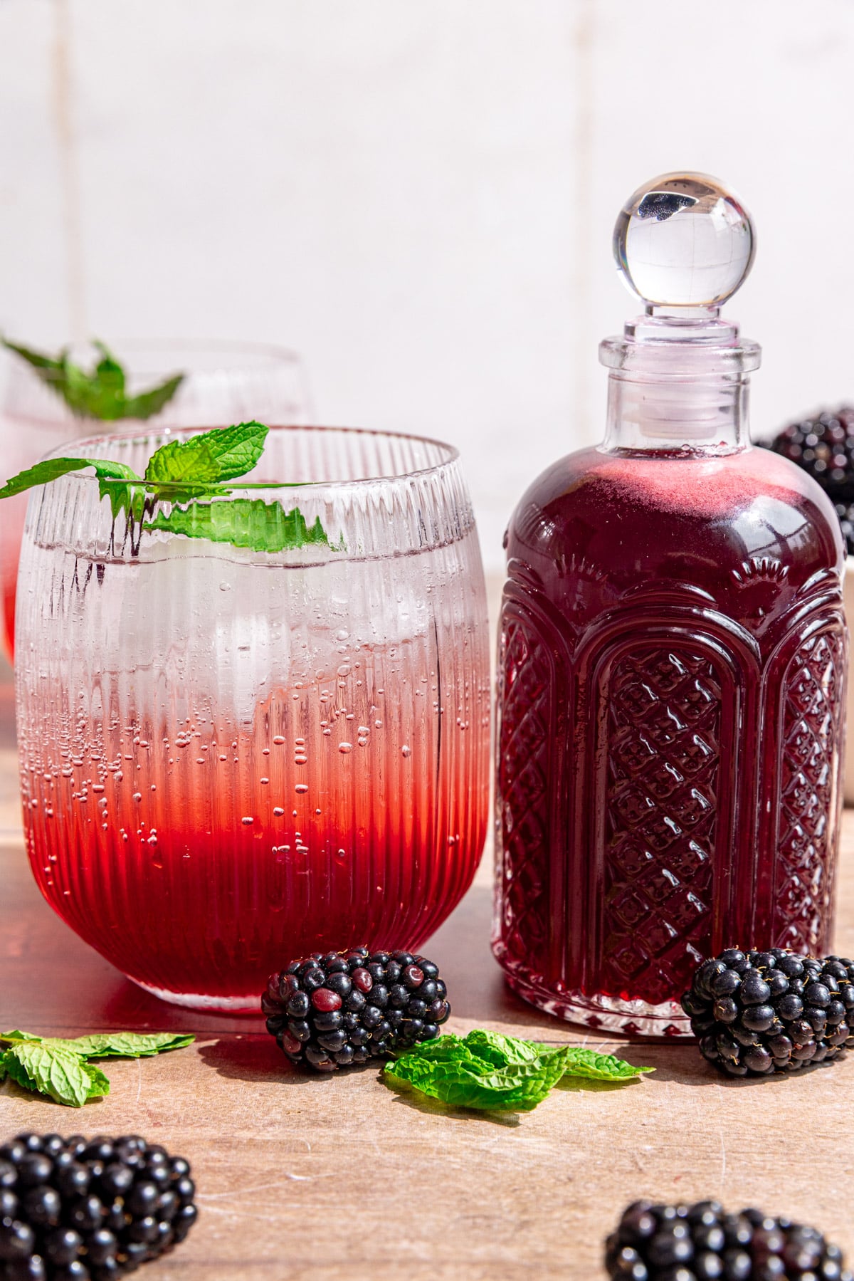 A bottle of blackberry shrub, next to a shrub mocktail and a berry basket of blackberries.