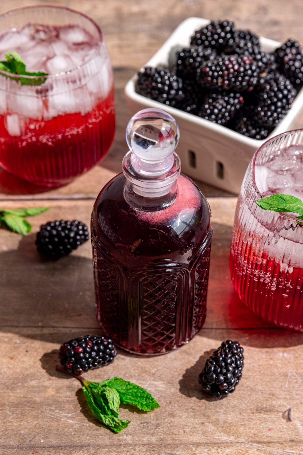 A bottle of blackberry shrub, next to a shrub mocktail and a berry basket of blackberries.