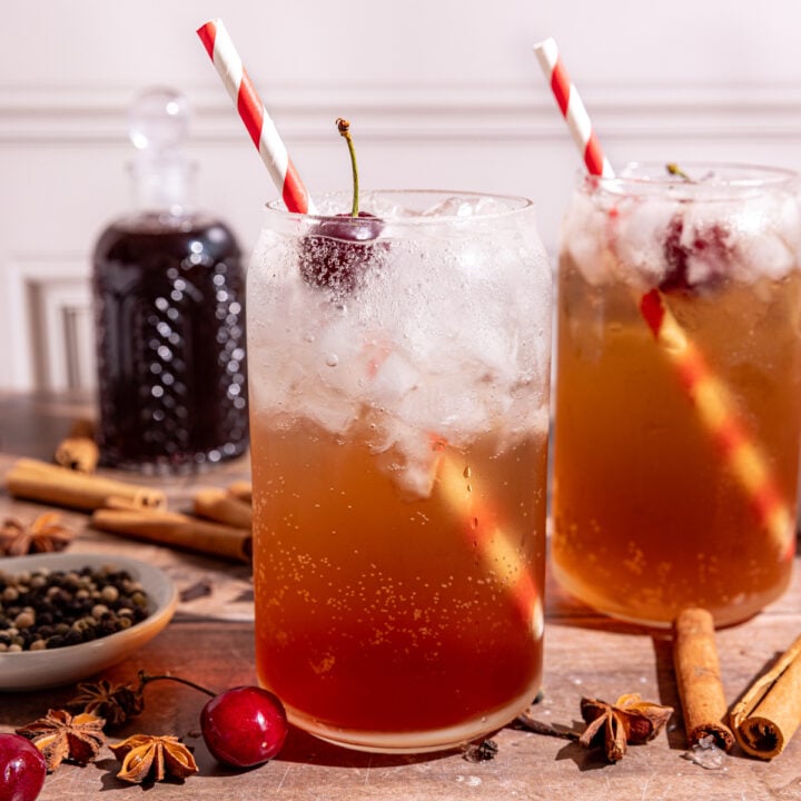 A soda glass of homemade Dr. Pepper with a cherry garnish and red & white striped straw.
