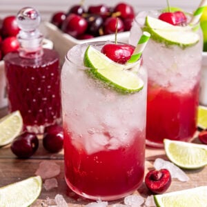Several glasses of cherry limeade soda, garnished with a fresh cherry, lime wedge, and green and white striped straw.