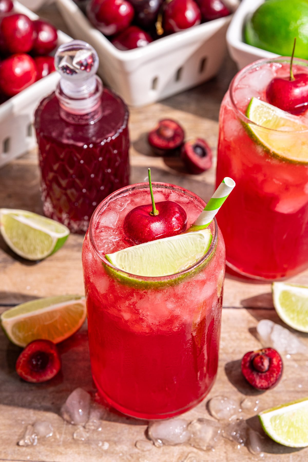 Several glasses of cherry limeade soda, garnished with a fresh cherry, lime wedge, and green and white striped straw.