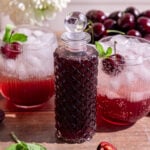 A bottle of cherry shrub, next to two cherry shrub mocktails.