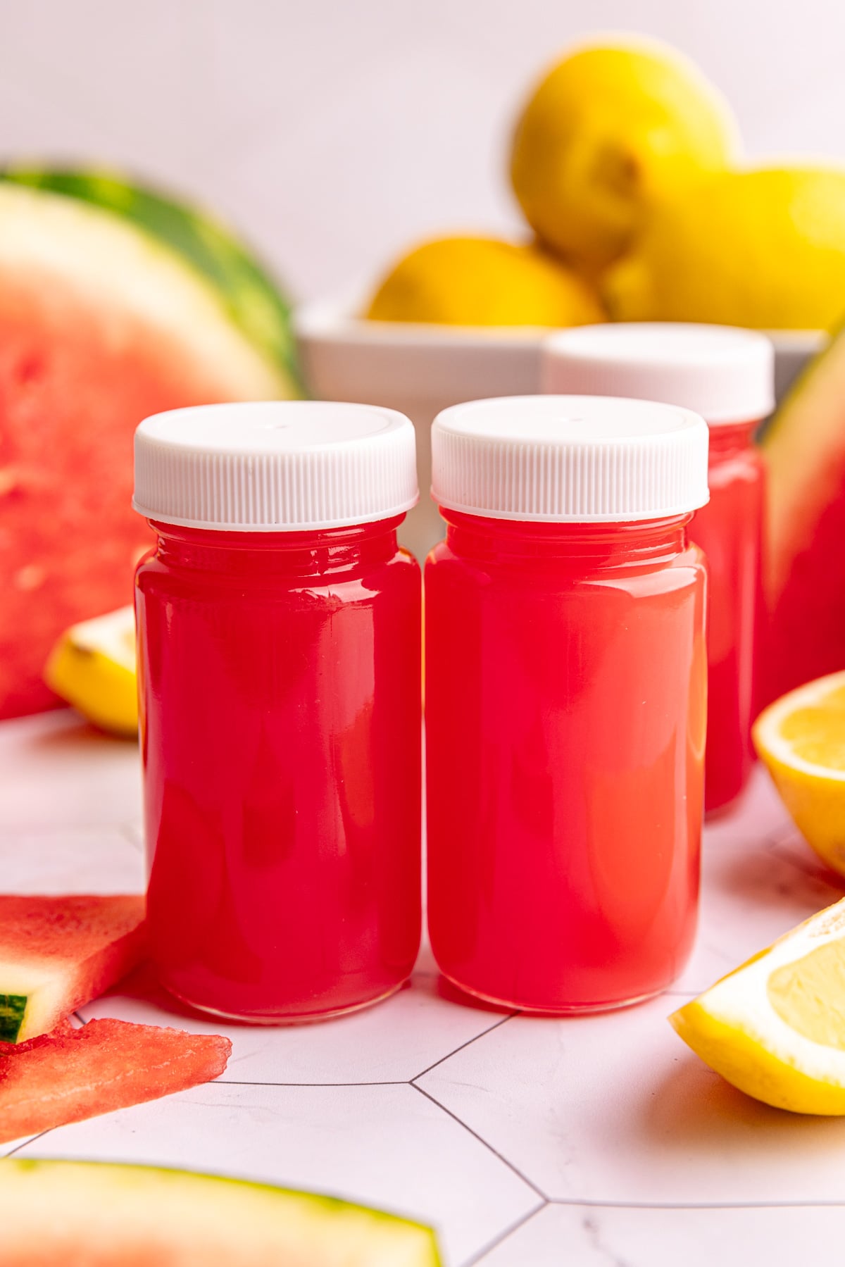 Watermelon ginger wellness shots on a countertop with fresh lemons and watermelon slices.