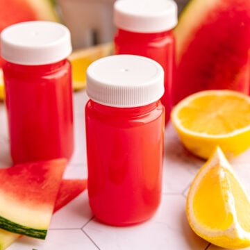 Watermelon ginger wellness shots on a countertop with fresh lemons and watermelon slices.